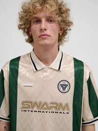 Ung mann med krøllete blondt hår iført en beige og grønn poloskjorte med "SWARM INTERNATIONALS"-logo og gull hoop-øredobber, nøytralt uttrykk.