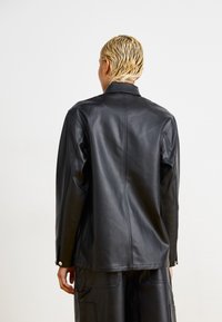 Giacca in pelle nera con una texture liscia, dotata di una vestibilità su misura, colletto a punta e dettagli con bottoni a pressione sulle maniche.