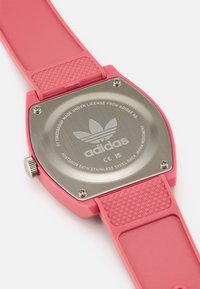 Rozā plastmasas pulkstenis ar kvadrātveida nerūsējošā tērauda aizmuguri, struktūras silikona siksniņu un izgravētu Adidas logo. Iekļauj arī kroni sānos.