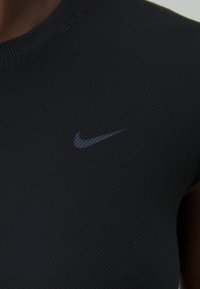 Črna rebrasta športna majica z okroglo ovratnikom, z nevsiljivim črnim logotipom Nike na sprednji strani. Gladka tekstura, prilagojen dizajn.