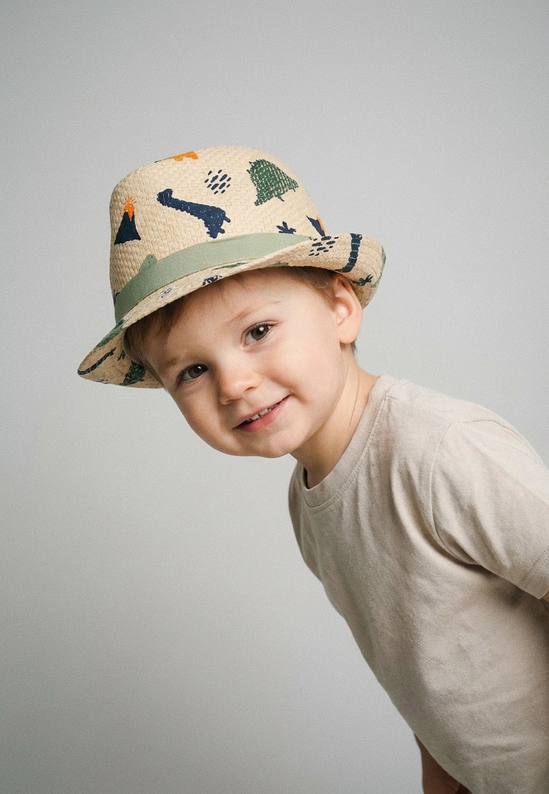 Jeune garçon aux cheveux châtain clair portant un t-shirt beige et un chapeau de paille à motifs, souriant et penché en avant sur un fond uni.