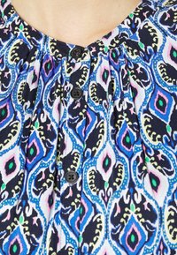 La tela de múltiples colores presenta un fondo azul marino con diseños en púrpura, verde y rosa. Cuatro botones negros adornan la parte delantera.
