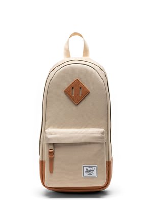 Béžový batoh s predným vreckom na zips, hnedým koženým dnom a logom na nášivke, a koženou nášivkou v tvare diamantu na hornej prednej časti.