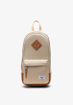 Mochila beige con bolsillo delantero con cremallera, base de cuero marrón y parche con logo, y un parche de cuero en forma de diamante en la parte superior delantera.