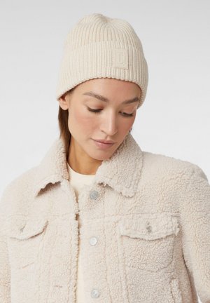 Beige knit beanie with a ribbed texture and a small logo patch. Paired with a light cream, fluffy shearling jacket.