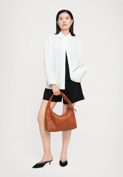Sac à main tissé marron avec une forme arrondie et une surface texturée ; exposé avec une chemise claire, un short noir et des mules à talons noirs.