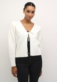 Cardigan en maille blanche avec un motif en losange, décolleté en V, fermeture à l'avant par un lien, et ourlet côtelé. Porté par-dessus un haut noir et associé à un jean foncé.