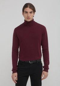 Giovane uomo con capelli castani di media lunghezza, che indossa un maglione attillato bordeaux e pantaloni neri con una cintura nera, in piedi contro uno sfondo semplice.