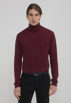 Giovane uomo con capelli castani di media lunghezza, che indossa un maglione attillato bordeaux e pantaloni neri con una cintura nera, in piedi contro uno sfondo semplice.