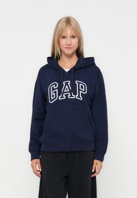 Marinblå zip-up huvtröja i mjukt tyg med en stor vit "GAP" logotyp på framsidan, samt snörning i huvan och framfickor.