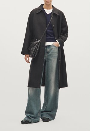 Schwarzer Wollmantel mit großen Revers, darüber ein marineblauer Pullover und ein weißes Unterhemd, kombiniert mit lockeren hellblauen Jeans und schwarzen Schuhen.