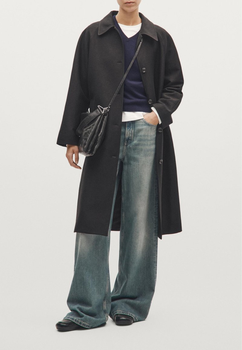 Černý vlněný kabát s velkými klopami, nošený přes námořnický svetr a bílou košili, doplněný o volné světle modré džíny a černé boty.