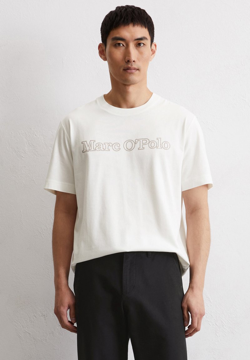Weißes Baumwoll-T-Shirt mit einem vorderen Logo "Marc O'Polo" in einer gedämpften Farbe. Kurzärmelig und mit Rundhalsausschnitt, kombiniert mit dunklen Hosen.