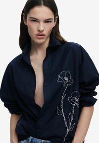 Navyblaues Hemd mit Knöpfen, tiefem V-Ausschnitt und langen Ärmeln, mit weißer floralem Stickerei auf der linken Seite. Glatte Stofftextur.