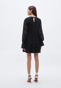 Μαύρο φόρεμα από δαντέλα με μακριά μανίκια, φαρδιά μανσέτα και κυκλικό στρίφωμα. Διαθέτει άνοιγμα σε σχήμα κλειδιού στην πλάτη. Φοριέται με κόκκινα ψηλοτάκουνα και λευκές κάλτσες.