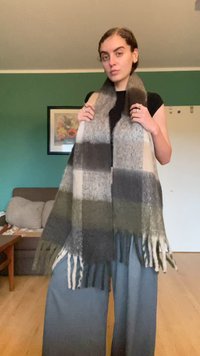 Szürke, olívazöld és krém színű kockás mintázatú gyapjú sál, bojtolt szélekkel és puha textúrával, a modell vállán drapírozva.