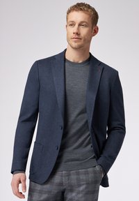 Blazer navy a trama con rever, due bottoni e taschino sul petto, abbinato a una maglietta grigia con collo rotondo e pantaloni con fantasia.