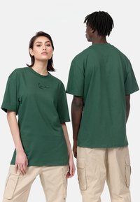 T-shirt en coton vert à manches courtes, avec un motif graphique noir sur le devant. Coupe ample, col rond, dos uni, porté avec un pantalon cargo beige.
