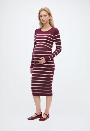 Femme enceinte portant une robe ajustée à manches longues rayée bordeaux et blanche, avec des ballerines bordeaux, debout avec une main sur son ventre.