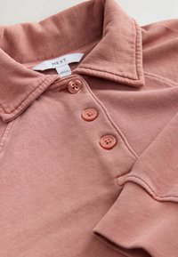 Jasnoróżowa koszulka polo wykonana z miękkiego materiału, z kołnierzykiem, trzema guzikami i teksturowanymi mankietami. Metka mówi "NEXT".