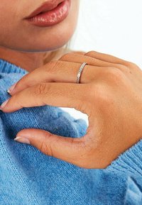 Dünner Silberring mit kleinen klaren Steinen, getragen an einer Hand, die einen blauen Pullover hält. Der Pullover hat ein weiches, strukturiertes Aussehen.