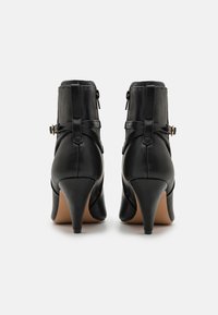 Botas de tobillo de cuero negro con punta afilada, tacón alto y estrecho, y cremalleras laterales. Presentan correas decorativas y acentos metálicos.