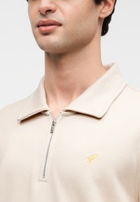 Junger Mann trägt ein beiges Reißverschlusshemd mit Kragen und einem kleinen gelben, gestickten Vogel auf der linken Brust.