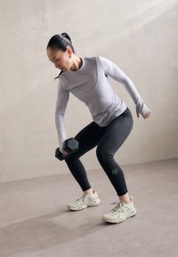 Kvinna i träningskläder som utför en stående hantelrodd i ett minimalistiskt inomhusrum.