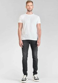 Camiseta blanca de manga corta combinada con jeans oscuros ajustados y zapatillas en blanco y negro, de pie contra un fondo neutro.