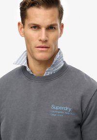 Szary sweter z okrągłym, prążkowanym dekoltem, z niebieskim logo Superdry i napisami miast nadrukowanymi na przodzie. Pod spodem widoczny jasnoniebieski kołnierzyk w paski.