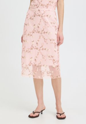 Różowa przezroczysta spódnica midi z haftem kwiatowym, noszona z brązowymi sandałami na obcasie na nogach na białym tle.