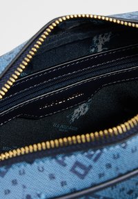 Wnętrze torby w niebieskie wzory z wieloma kieszeniami na zamek i gładkim, tekstylnym wykończeniem. Posiada złote elementy zamków i ciemną wyściółkę.