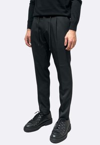 Pantaloni neri su misura con vita con coulisse, due pieghe frontali e tessuto liscio. Abbinati a scarpe nere stringate.