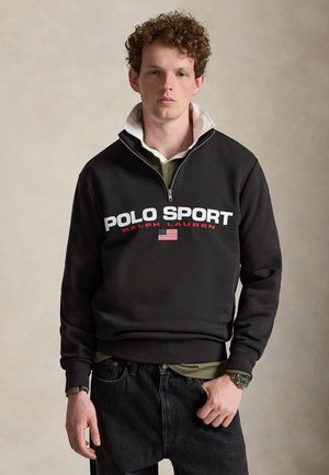 Jeune homme aux cheveux roux bouclés portant un sweat-shirt noir Polo Sport Ralph Lauren avec un col blanc et un emblème du drapeau américain.