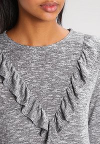 Šedý svetr s texturovanou tkaninou, který má výstřih do V a volánkový akcent podél výstřihu, zdůrazňující ležérní design.