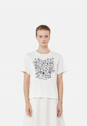Donna che indossa una T-shirt bianca con motivo floreale nero e la scritta "WEEKEND", abbinata a una gonna bianca su uno sfondo semplice.