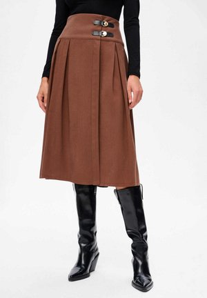 Jupe midi plissée marron taille haute avec deux ceintures noires à boucle, portée avec un haut noir à manches longues et des bottes en cuir noires jusqu'aux genoux.