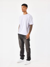 Camiseta corta de manga morada clara con logo bordado, combinada con jeans gris oscuro y zapatillas blancas. Fondo liso.