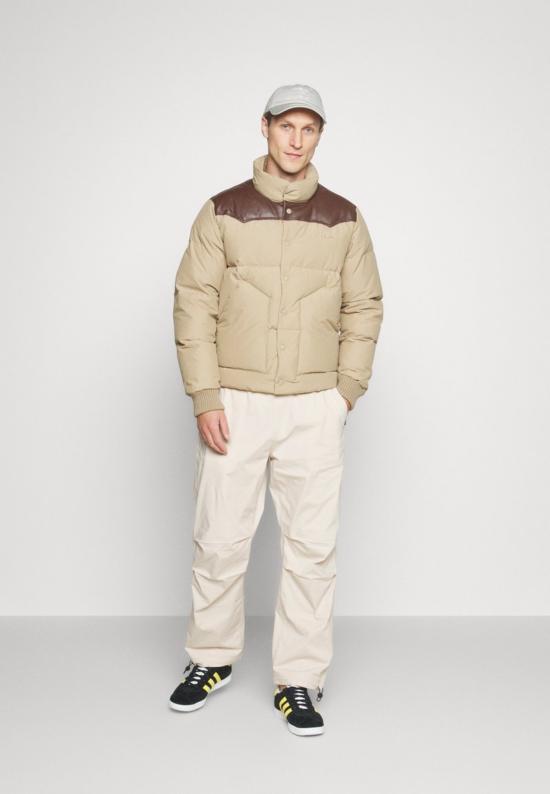 Uomo che indossa un piumino beige con dettagli marroni sulle spalle, pantaloni beige chiaro, sneaker nere con strisce gialle e cappellino grigio chiaro in piedi.