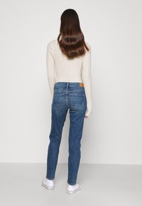 Těsné tmavě modré džíny se žebrovaným béžovým svetrem. Džíny mají design zadní kapsy a hnědou koženou etiketu značky na pase.