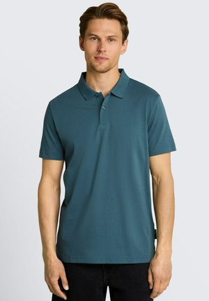 Jeune homme portant un polo manches courtes couleur sarcelle et un pantalon noir, debout devant un fond gris clair uni.