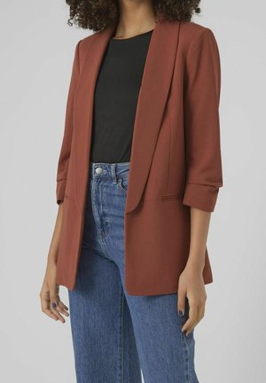 Femme portant un blazer couleur rouille à manches retroussées, un haut noir et un jean bleu taille haute sur un fond clair uni.