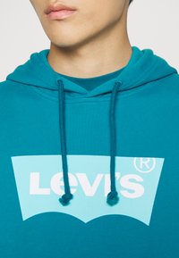 Teal capuchontrui met een zachte textuur. Groot lichtblauw Levi's logo op de voorkant, met een klassiek gebogen ontwerp. Verstelbare koorden.