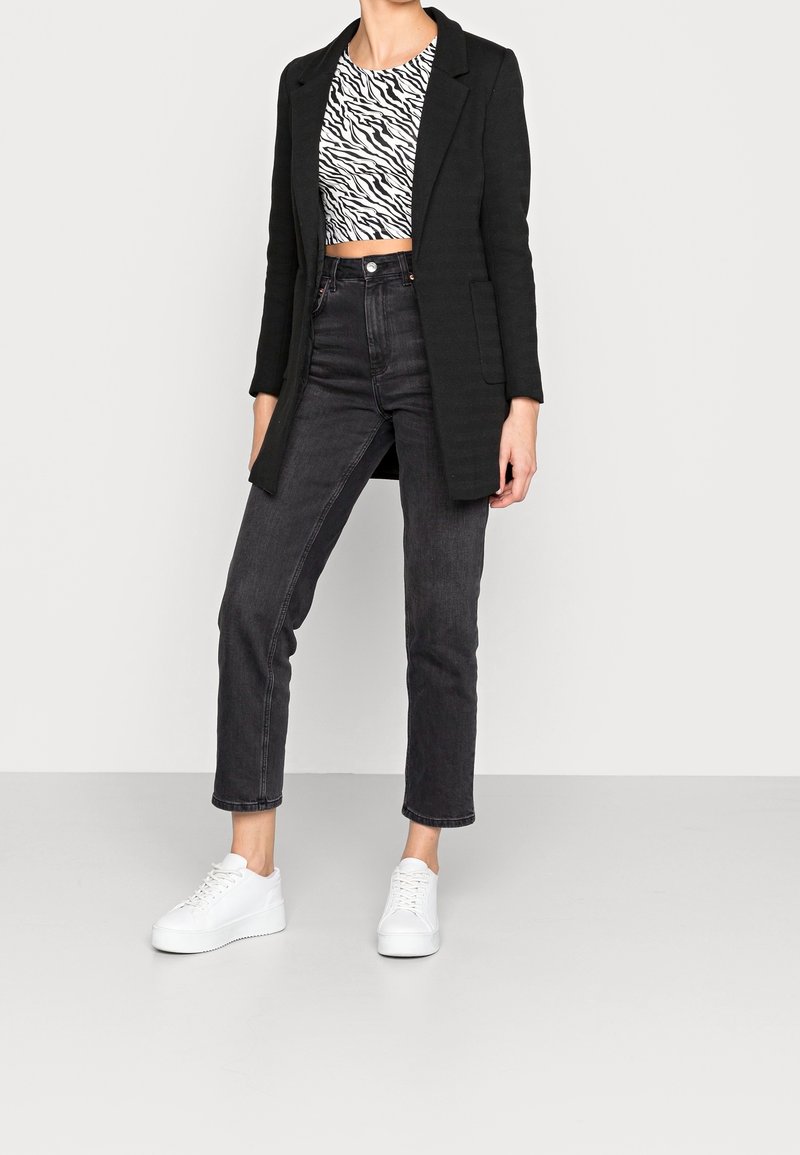 Blazer lungo nero sopra un crop top zebrato bianco e nero, abbinato a jeans neri a vita alta e sneakers bianche.
