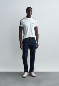 Modèle masculin debout portant un t-shirt blanc avec des rayures colorées sur les épaules, un jean bleu foncé, une ceinture noire et des baskets blanches.
