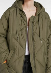 Olivgrön vadderad jacka med dragkedja fram, diamantquiltning och huva. Har silverfärgade detaljer och en innerficka med orange foder.