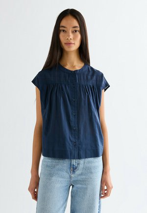 Femme aux longs cheveux noirs portant un chemisier sans manches bleu marine à boutons et un jean taille haute bleu clair sur un fond uni.