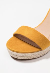 Žluté semišové sandály s širokým páskem a platformou z pleteného jutového materiálu. Podešev je béžová, s hladkým povrchem a zaobleným tvarem.