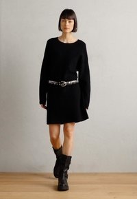 Vestido negro de punto con un corte holgado, mangas largas y bajo acampanado, acentuado por un cinturón metálico con tachuelas. La modelo lleva botas negras hasta medio pantorrilla.