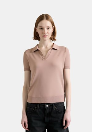 Giovane donna con capelli lisci, corti e castano chiaro che indossa una maglietta rosa polvere a maniche corte con collo e pantaloni neri su uno sfondo semplice.